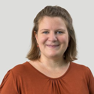 Eine junge Frau mit dunkelblonden Haaren und einer braunen Bluse vor einem grauen Hintergrund