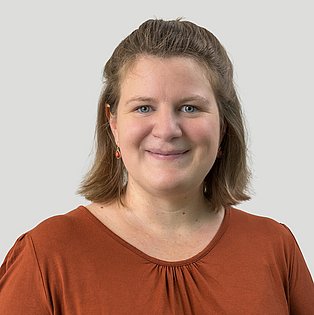 Eine Frau mit dunkelblondem Haar und einer braunen Bluse vor einem uni hellgrauen Hintergrund