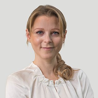 Eine Frau mit blondem Haar und gekleidet mit einer weißen Bluse vor einer hellgrauen Wand